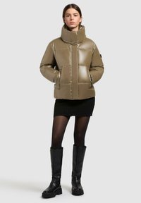 Giacca puffer khaki con colletto alto, motivo trapuntato, tasche con cerniera e finitura lucente, indossata sopra una gonna nera a coste e stivali neri alti.