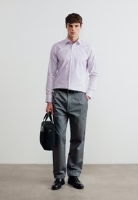 Άνδρας με ελαφρώς μωβ πουκάμισο και γκρι παντελόνι, κρατώντας μαύρη τσάντα στο αριστερό χέρι, στέκεται μπροστά σε λευκό φόντο.