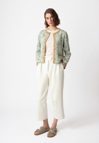 Zeleni s uzorkom sako s narančastim detaljima, nosi se preko lagane bluze i bijelih širokih hlača, u kombinaciji s bež sandalama.