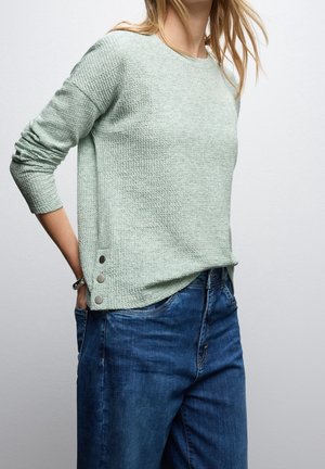 Femme portant un pull texturé vert clair à manches longues avec trois boutons sur le côté et un jean en denim bleu, main dans la poche.