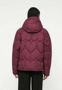 Giacca trapuntata invernale color bordeaux con imbottitura a forma di cuore, cappuccio e spalle scese. Tessuto morbido e strutturato con una vestibilità rilassata.