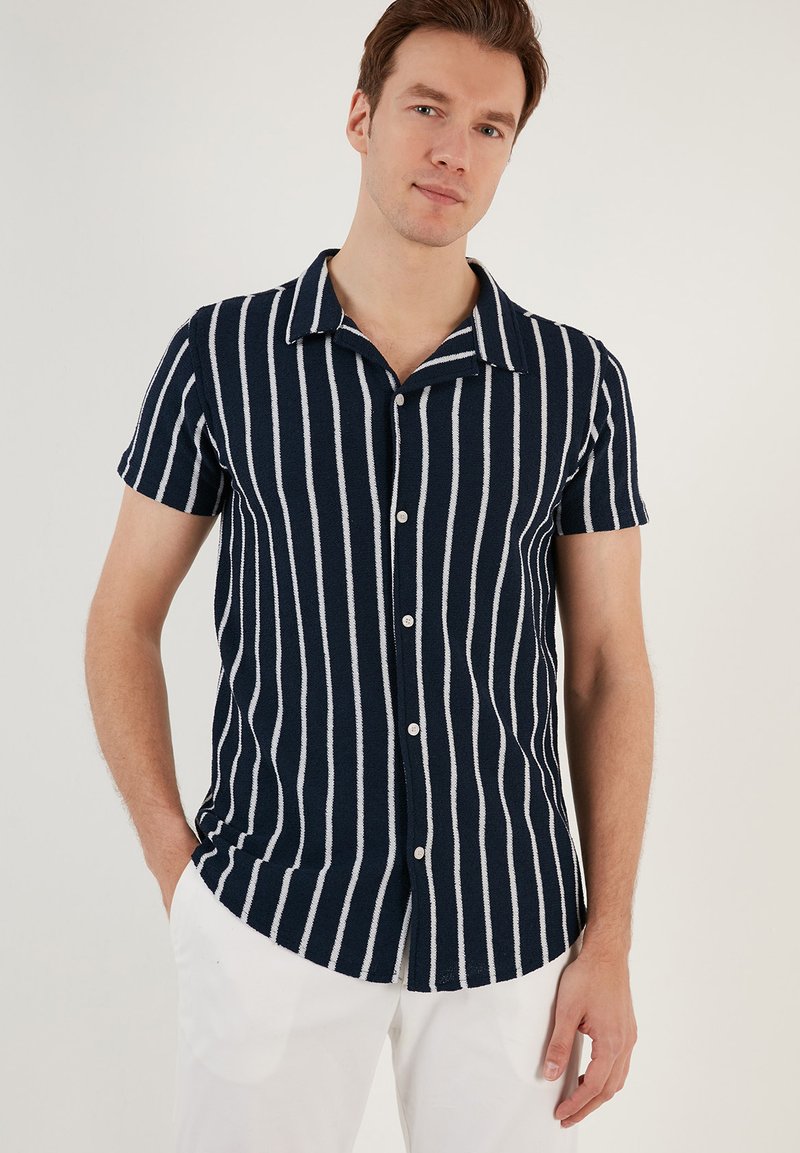 Camicia a maniche corte, con bottoni, di colore blu navy con strisce verticali bianche. Presenta un colletto a camicia e un tessuto texture.