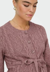 Aunāds džemperis mauve krāsā ar aušanas rakstu, apaļu kakla izgriezumu, trim priekšējiem pogām un sasaistītu vidukļa detaļu. Mīksta tekstūra ar ribotām akcentiem.