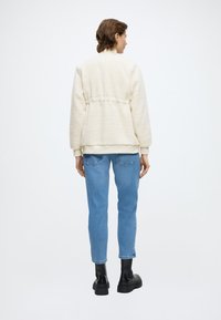 Giacca in pile color crema con superficie strutturata, vita aderente e polsini a costine, abbinata a jeans azzurri e stivali neri massicci.