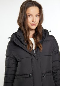 Jachetă puffer neagră cu glugă, având închidere cu fermoar și șnururi ajustabile, textură matlasată și un design ușor unghiular.