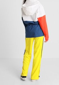 Blockfarbige Jacke mit weißen, dunkelblauen und roten Abschnitten; gelbe Trainingshose mit schwarzen Seitenstreifen; Schuhe mit niedrigem Profil.