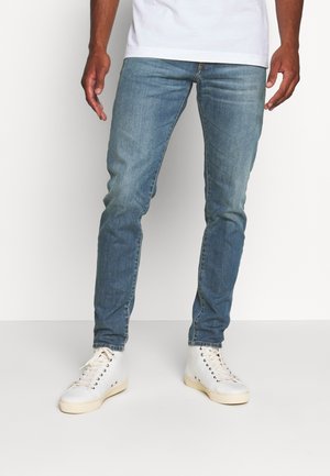 Homme portant un jean bleu slim et des baskets blanches montantes, debout devant un fond clair uni.