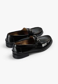 Mocasines de cuero negro con un acabado brillante, que presentan un detalle de costura elevada y una hebilla decorativa en la parte frontal. Suela plana de goma.