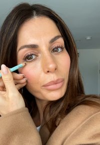 Una donna che applica una matita per eyeliner azzurro chiaro sulla palpebra inferiore, indossando un cappotto beige, con lunghi capelli castano scuro e trucco neutro.