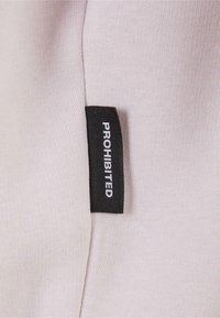 Tissu rose clair avec une surface texturée. Une étiquette noire indique "INTERDIT" en lettres blanches, fixée au bord du vêtement.