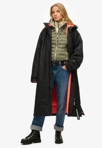 Abrigo negro largo e impermeable con forro naranja, superpuesto a una chaqueta acolchada verde claro. Jeans de mezclilla azules y botas negras con cordones completan el look.