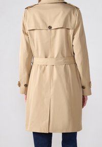 Beżowy płaszcz trench z paskiem w talii, wentylacją z tyłu, naramiennikami i guzikami na rękawach. Gładka, strukturalna tekstura materiału.