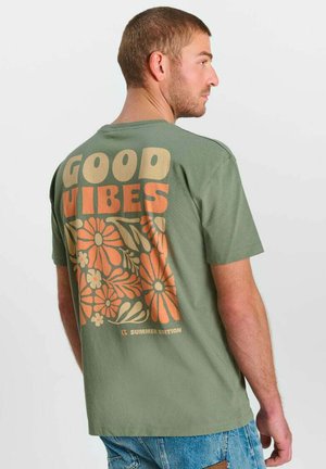 Uomo con capelli corti che indossa una maglietta verde con motivo floreale arancione e la scritta "Good Vibes" sul retro, abbinata a un paio di jeans blu.