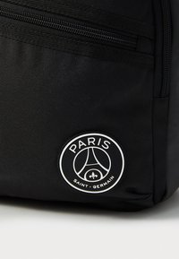 Fekete hátizsák tartós szövetből, cipzáras zsebbel és egy kerek logóval, amelyen fehér színnel szerepel a "PARIS" és "SAINT-GERMAIN" felirat.