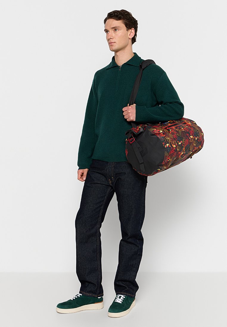 Uomo che indossa un maglione verde scuro, jeans scuri, sneaker verdi e porta una grande borsa a tracolla con motivo floreale.