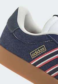 Navy denim sneaker with a rubber gum sole, featuring white and red stripes, textured accents, and a suede heel patch. Gold logo detail present.