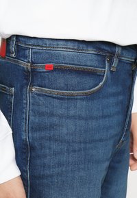 Jeans in denim blu con una vestibilità slim, cuciture a contrasto, tasche frontali e un'etichetta del marchio rossa vicino alla vita.