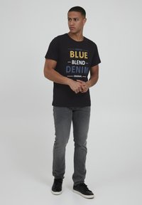 Melnās kokvilnas T-krekls ar krāsainu tekstu: "VINTAGE BLUE BLEND DENIM ORIGINAL," kombinēts ar pelēkām džinsām un melnā sporta apaviem.