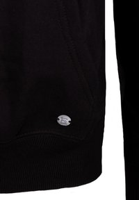 Sudadera negra con una textura suave, costuras en ángulo y una etiqueta de logo metálica. El dobladillo tiene detalles acanalados para mayor estructura.