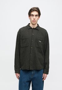 Camisa às riscas em preto e verde, com botões e colarinho, apresentando um bolso e um discreto logo branco. Combinada com jeans de ganga azuis.