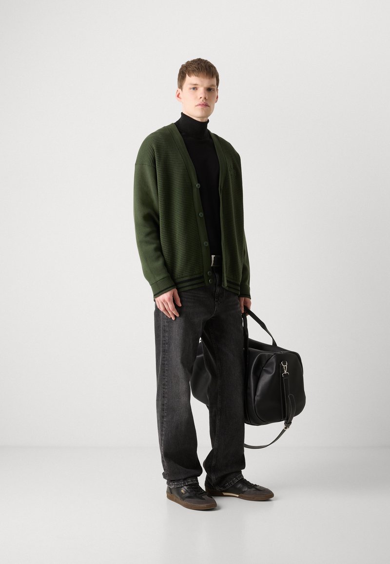 Zielony żebrowany kardigan, czarny golf, ciemne dżinsy, szare buty. Nosi dużą czarną skórzaną torbę z podwójnymi uchwytami i paskiem na ramię.