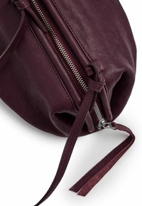 Bolso de cuero burdeos con forma redondeada, detalle de cremallera plateada y dos largas asas de cuero. Textura suave con una apariencia ligeramente arrugada.