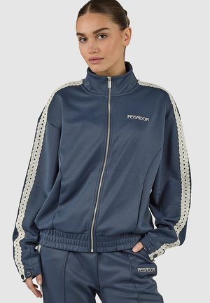 Young woman wearing navy blue tracksuit with white lace stripes on sleeves and pants, front zipper, and hands in pockets.