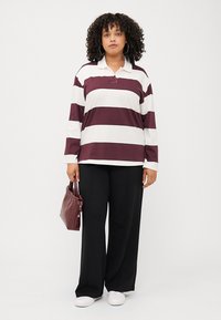 Une femme aux cheveux bouclés porte une chemise à rayures à manches longues et un pantalon noir à jambes larges, tenant un sac à main bordeaux sur un fond blanc.