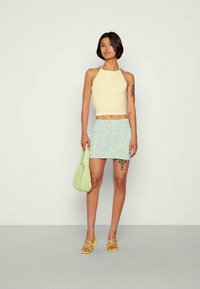 Mulher vestindo um top halter amarelo pálido, uma saia mini floral verde menta, sapatos de salto amarelos e segurando uma bolsa trançada verde clara, em pé contra um fundo neutro.