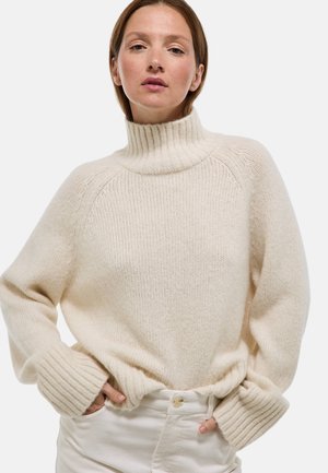 Un pull à col roulé de couleur crème en maille douce et texturée. Il présente des poignets côtelés et une coupe décontractée avec des épaules tombantes.