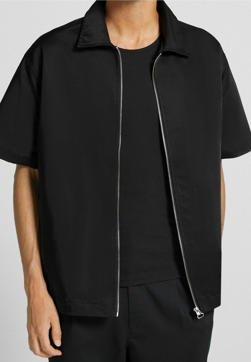Czarna krótka kurtka z kołnierzem typu stójka, z przednim zamkiem błyskawicznym i gładką fakturą, zestawiona z czarną koszulką z okrągłym dekoltem pod spodem.