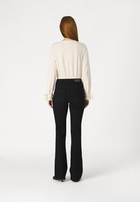 Femme aux longs cheveux roux portant un blouse crème à manches longues avec des poignets en ruban et un jean flare noir taille haute, se tenant de dos.