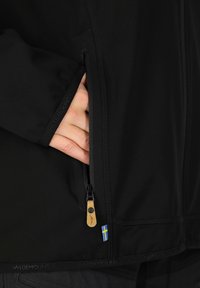 Svart softshell-jacka med ficka med dragkedja. Notabla detaljer inkluderar en blå och gul etikett och en läderdragkedjeanordning. Materialet verkar vara slätt.