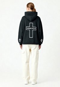 Felpa nera con cappuccio con la scritta "KARMA KILLS" in bianco e un design a croce bianca sul retro; abbinata a pantaloni cargo beige.