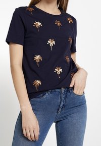 Námořnické bavlněné tričko s rozptýlenou bronzovou výšivkou palmy. Krátké rukávy, kulatý výstřih, uvolněný střih a měkký materiál.