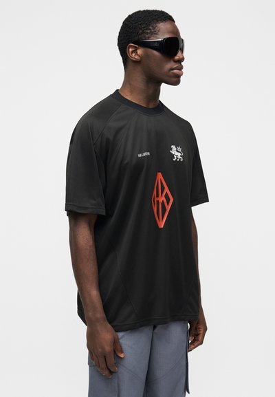 Black short-sleeve sports shirt with a round neck, featuring a white lion logo and red diamond graphic on the front. Smooth fabric texture.