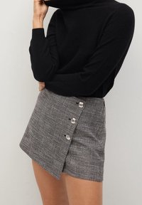 Jupe mini à carreaux gris texturé avec une coupe diagonale, ornée de boutons en argent sur le côté. Portée avec un pull col roulé noir.