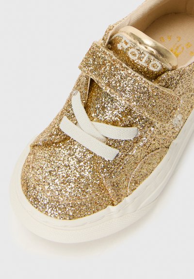 Sapatinho de criança dourado com glitter, cordões brancos, fecho de Velcro e sola branca de borracha sobre fundo branco.
