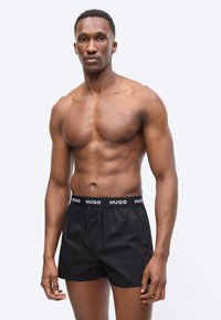 Μυώδης άνδρας όρθιος, φοράει μαύρα μπόξερ με το λογότυπο Hugo, κοιτώντας μπροστά με ουδέτερη έκφραση σε απλό ανοιχτό φόντο.