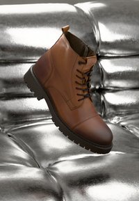 Bota de cuero marrón hasta el tobillo con diseño de cordones, suela de goma, puntera redondeada y detalles de costura, contra un fondo texturizado metálico.