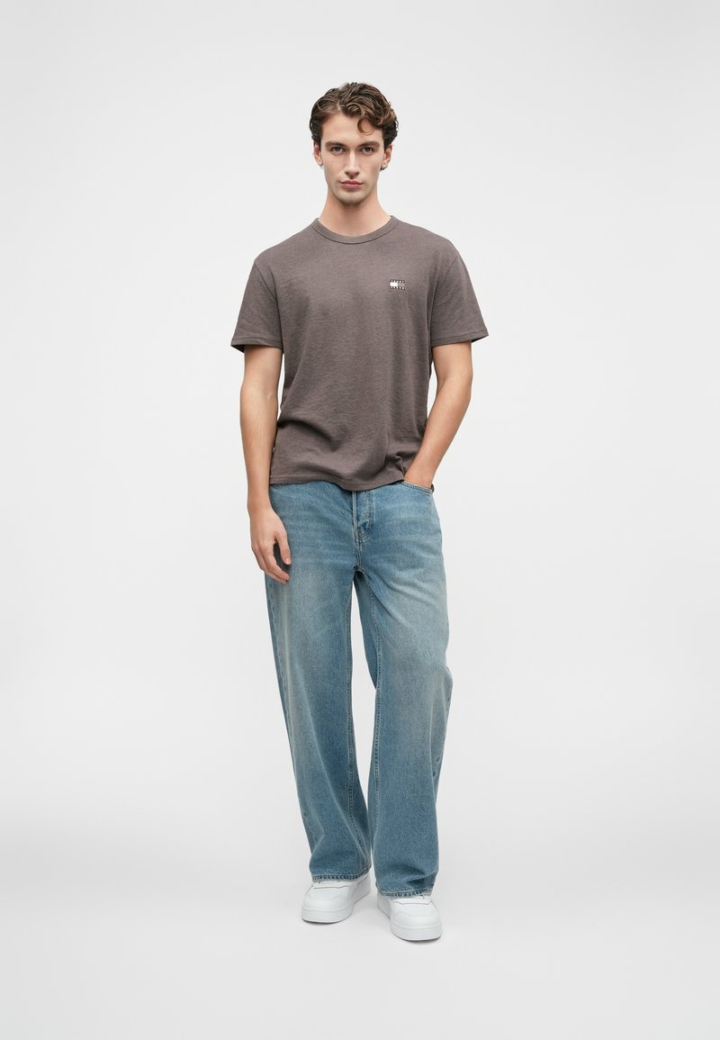 Ung mand med krøllet hår iført en brun t-shirt, brede lyseblå jeans og hvide sneakers, stående med den ene hånd i lommen.