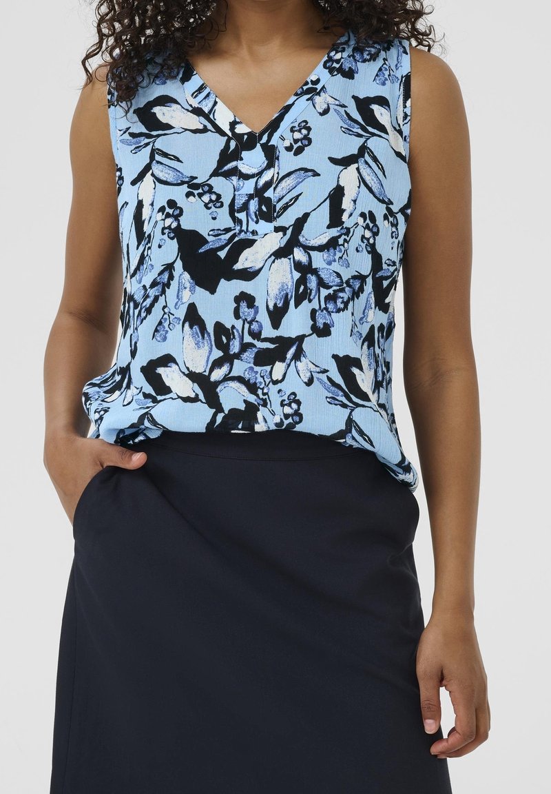 Femme portant un haut bleu à fleurs sans manches rentré dans une jupe bleu marine foncé, avec une main dans la poche de la jupe.