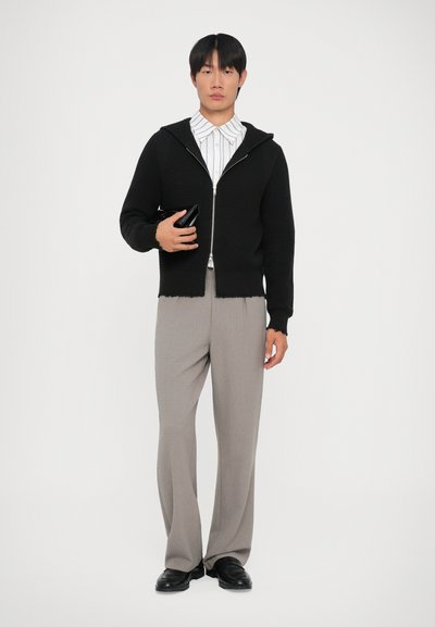 Svart stickad zip-up hoodie med ett texturerat mönster, bärs över en vit randig skjorta, kombinerad med ljusgrå byxor och svarta skor.