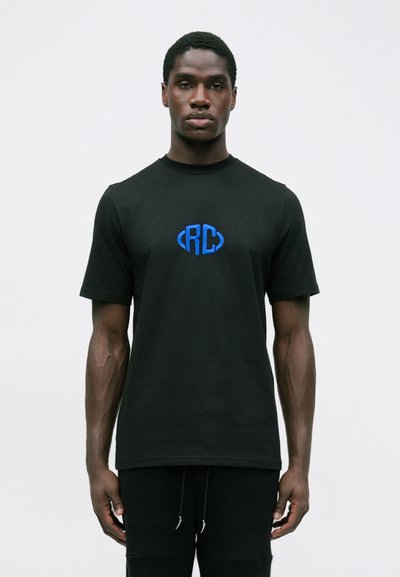 Tânăr cu pielea închisă la culoare, purtând un tricou negru cu un logo albastru "RC", stând pe un fundal alb simplu.