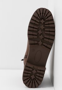 Calzado marrón con suela de goma, que presenta un patrón de relieve texturizado y un diseño de perfil bajo. La parte superior tiene un detalle de costura para mayor soporte.
