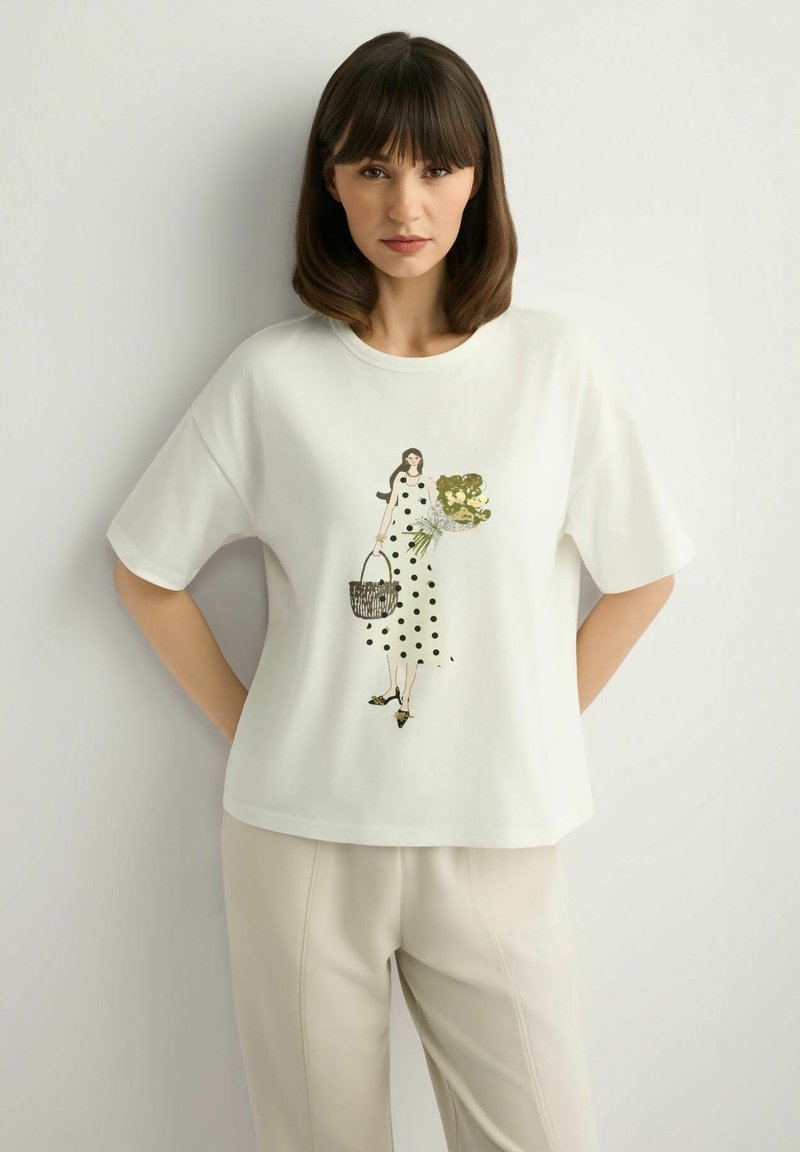 Kobieta o ciemnych, sięgających do ramion włosach nosi białą, oversize'ową koszulkę z ilustracją kobiety w sukience w groszki, trzymającej kwiaty i koszyk.