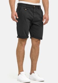 Shorts en coton noir avec revers roulés, poches avant et fermeture à un seul bouton. Portés avec des baskets blanches.