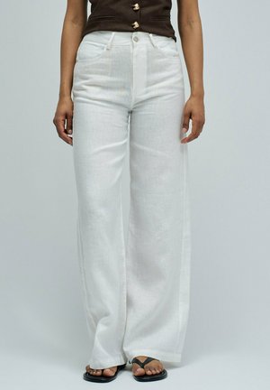 Persona con pantalones blancos de cintura alta y pierna ancha, top negro sin mangas con botones dorados y sandalias negras de playa, de pie frente a un fondo liso.