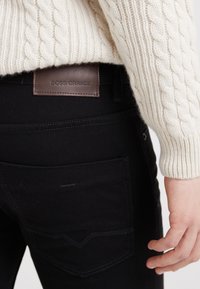 Black jeans with a brown leather "BOSS ORANGE" label, featuring a rear pocket with a curved stitching detail. Textured cream sweater partially visible.