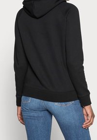 Sudadera negra con una capucha amplia y puños acanalados, combinada con vaqueros azules que presentan un diseño clásico de cinco bolsillos y un lavado desvaído.